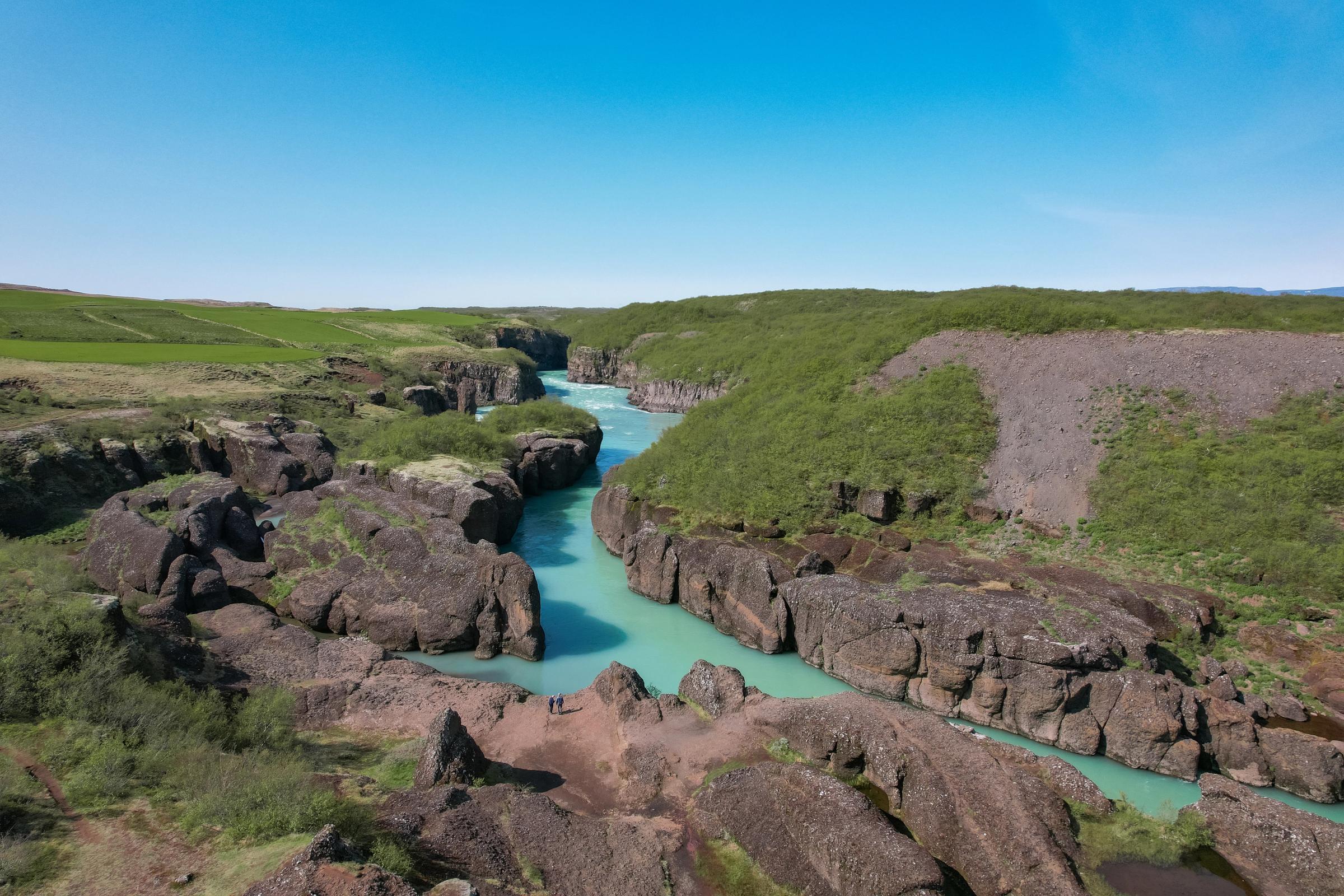 A turquoise river flows through a rocky canyon surrounded by green fields and hills under a clear blue sky.