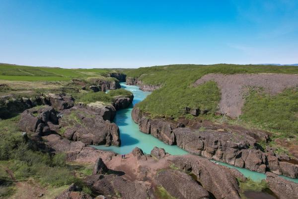 A turquoise river flows through a rocky canyon surrounded by green fields and hills under a clear blue sky.