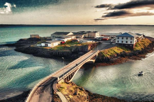 Painterly aerial view of a village on an island connected by a bridge over turquoise water, with a boat.