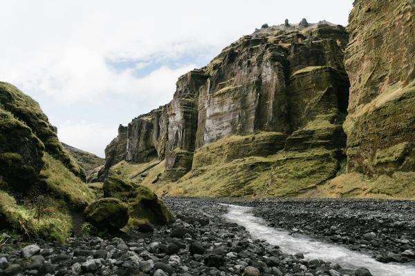 A river flows through a rocky canyon with steep, moss-covered walls under a cloudy sky.