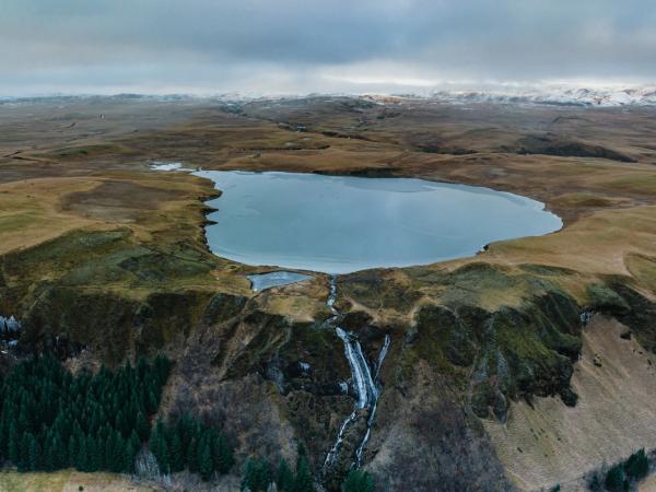 Aerial view of a lake feeding a waterfall that drops down a cliff face next to a small forest, surrounded by grassy hills.