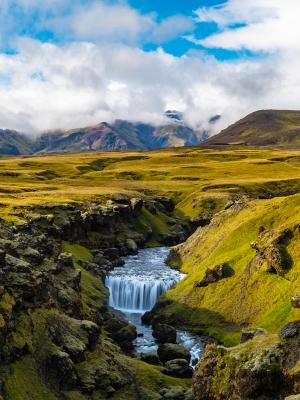 A waterfall in a rocky canyon surrounded by vibrant green and yellow hills with mountains under a partly cloudy sky.