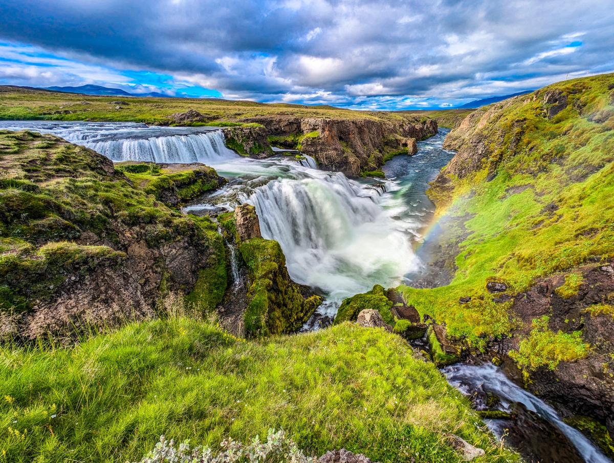 A powerful waterfall flows through a green, rocky canyon with a rainbow arching through the mist under a cloudy sky.