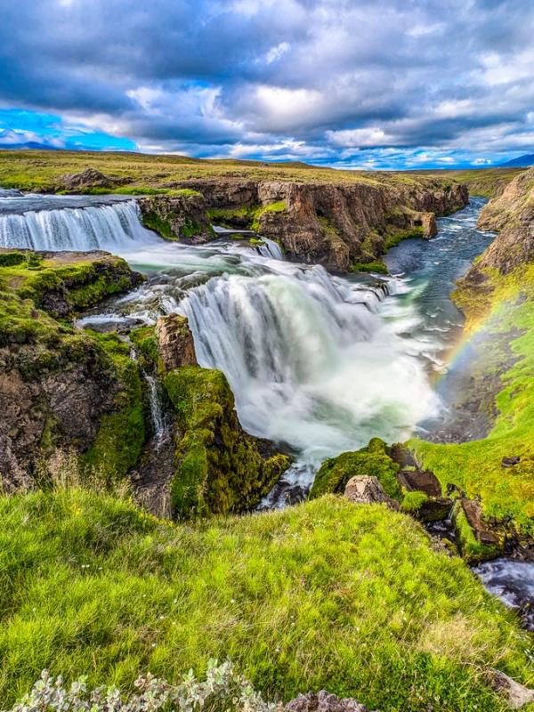 A powerful waterfall flows through a green, rocky canyon with a rainbow arching through the mist under a cloudy sky.