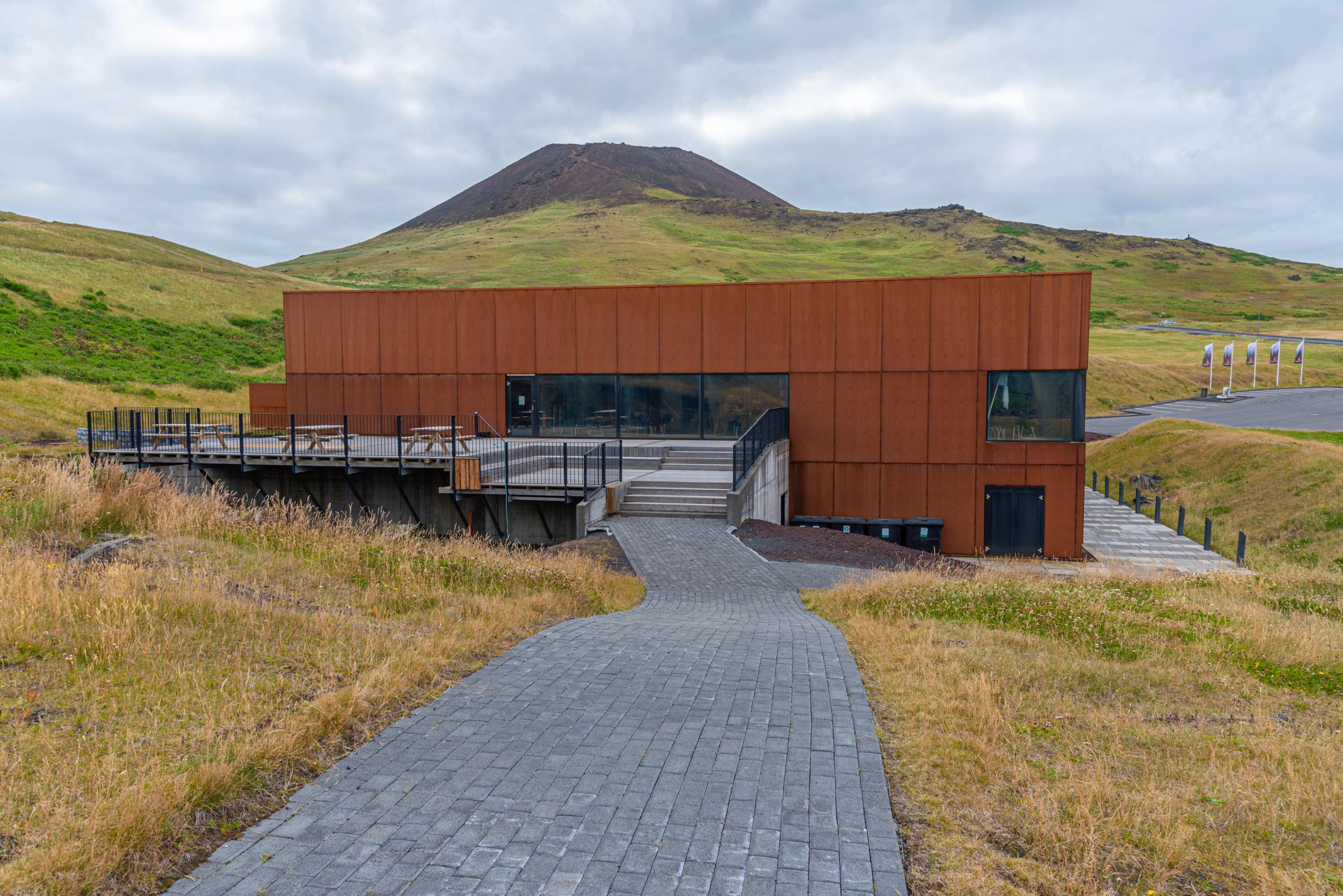 A modern, rust-colored building with a wooden deck in a grassy, hilly landscape with a volcanic cone in the background.