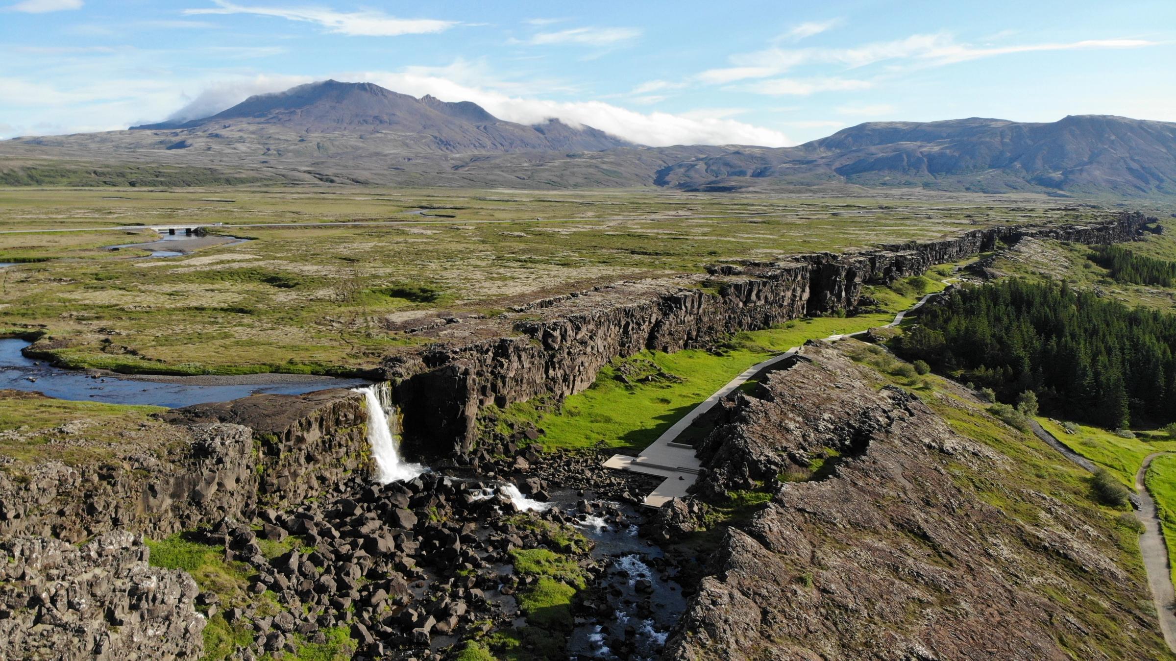 Una larga grieta rocosa con una cascada a la izquierda, que atraviesa una llanura verde con montañas distantes bajo un cielo azul.