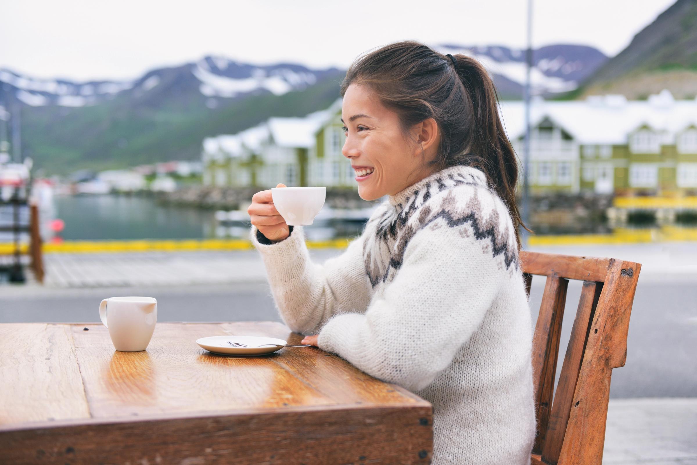 Mujer sonriente con un suéter estampado toma café en una mesa al aire libre con un puerto y montañas de fondo.
