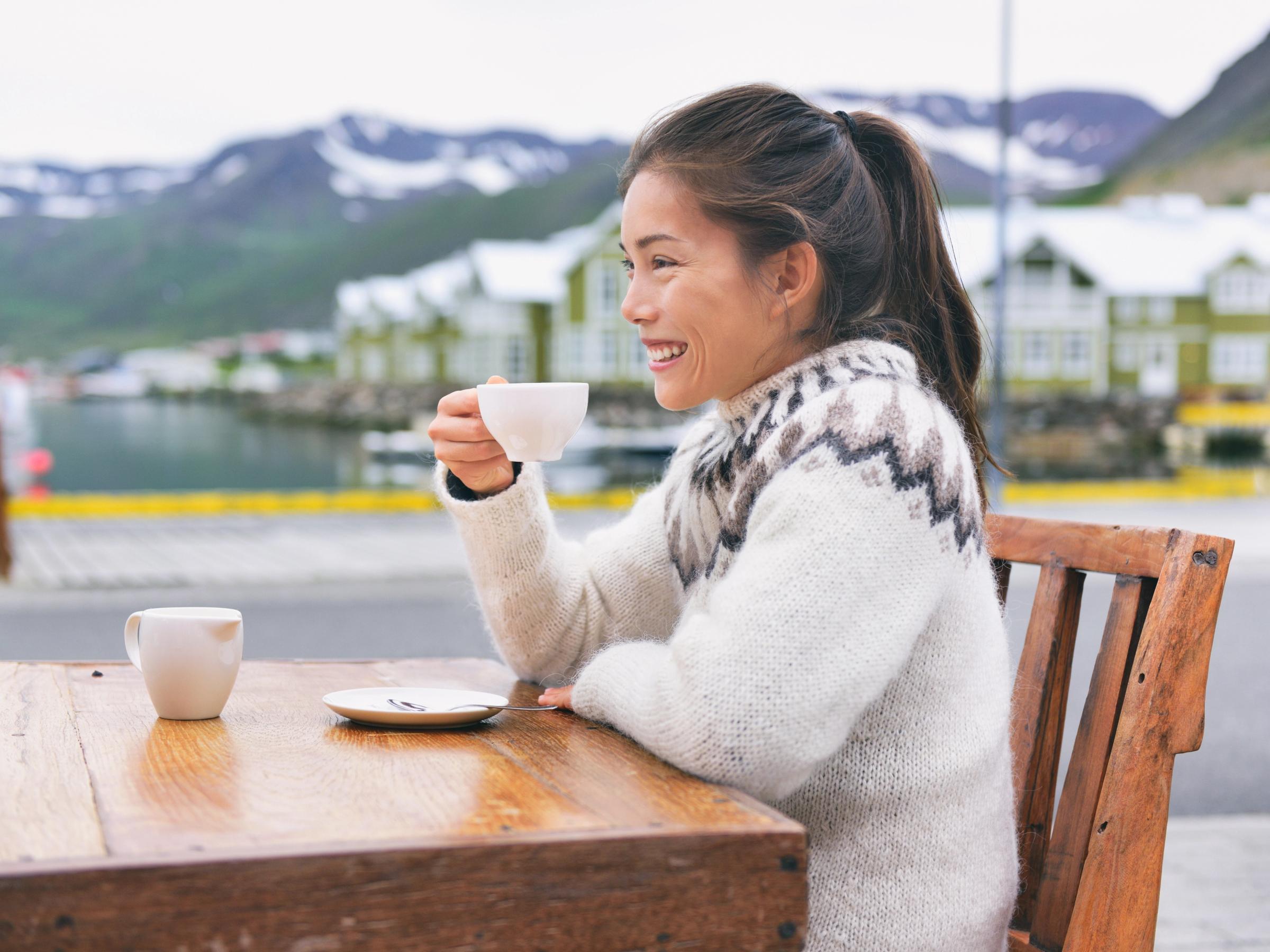 Mujer sonriente con un suéter estampado toma café en una mesa al aire libre con un puerto y montañas de fondo.