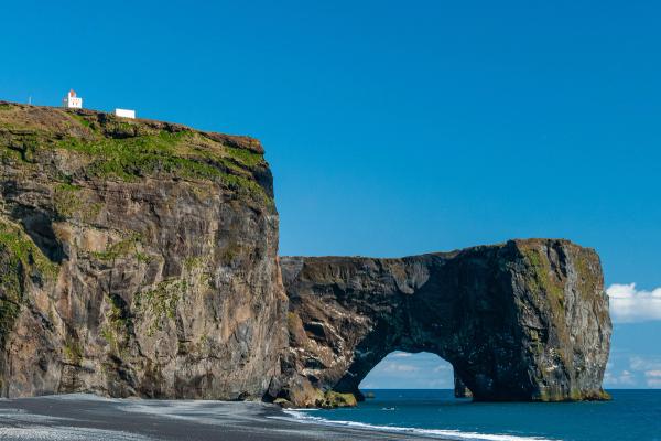 Dyrhólaey arch and black sand beach with a lighthouse on the cliff under a clear blue sky.