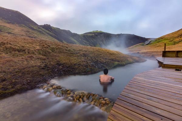 A person bathes in a steaming natural hot spring surrounded by hills, with a wooden deck to the right.