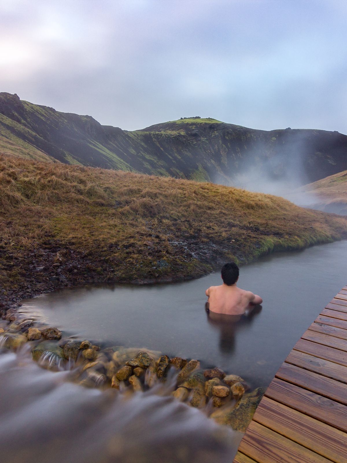 A person bathes in a steaming natural hot spring surrounded by hills, with a wooden deck to the right.