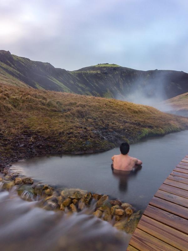 A person bathes in a steaming natural hot spring surrounded by hills, with a wooden deck to the right.