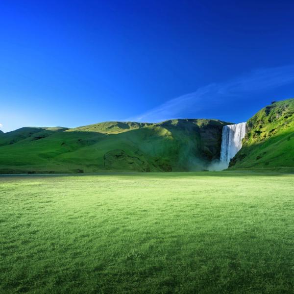 A lush green field with rolling hills and a tall waterfall under a vibrant blue sky.