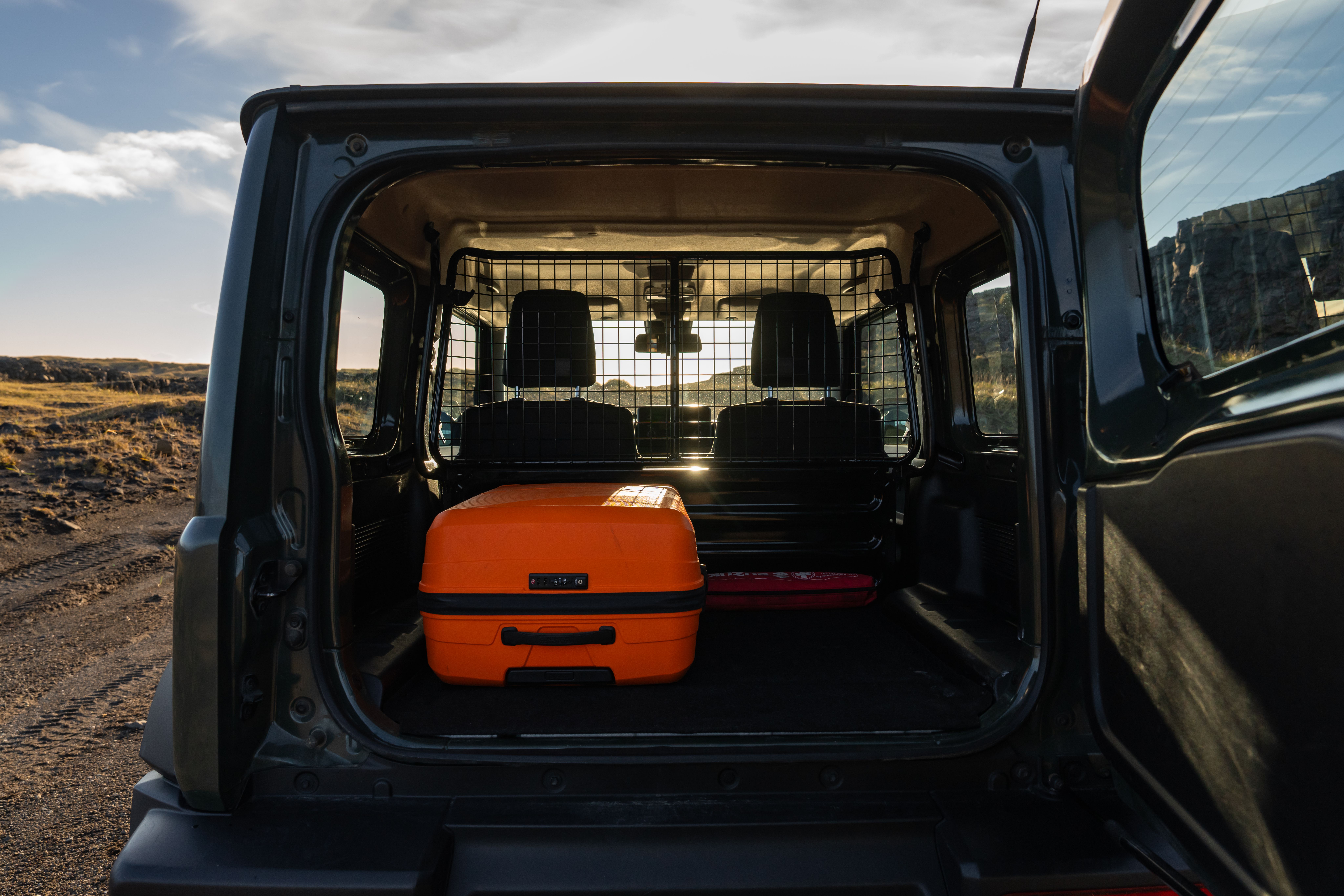 An orange suitcase and a red first aid kit sit in the open cargo area of a dark SUV, separated from the rear seats by a metal grid.