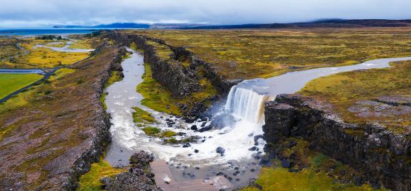 A river flows through a rocky fissure, forming a waterfall, surrounded by a vast vegetated plain under an overcast sky.
