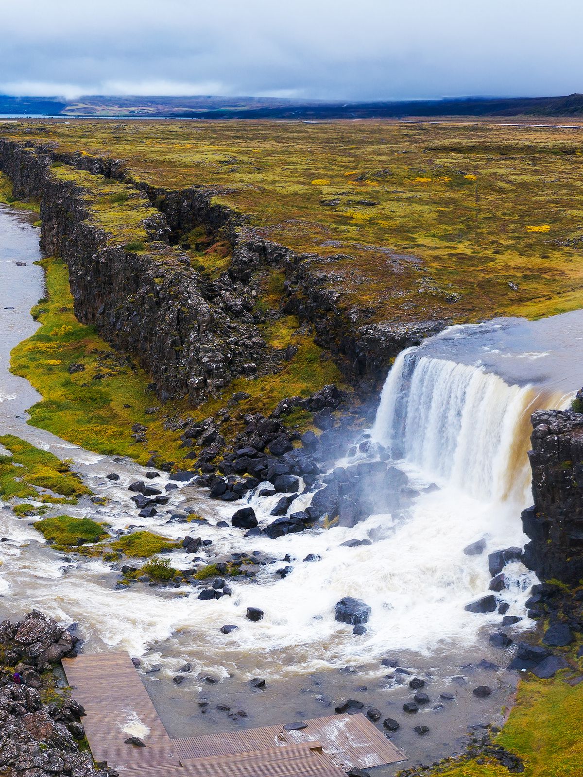 A river flows through a rocky fissure, forming a waterfall, surrounded by a vast vegetated plain under an overcast sky.
