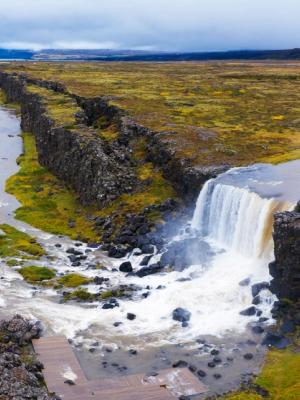 A river flows through a rocky fissure, forming a waterfall, surrounded by a vast vegetated plain under an overcast sky.