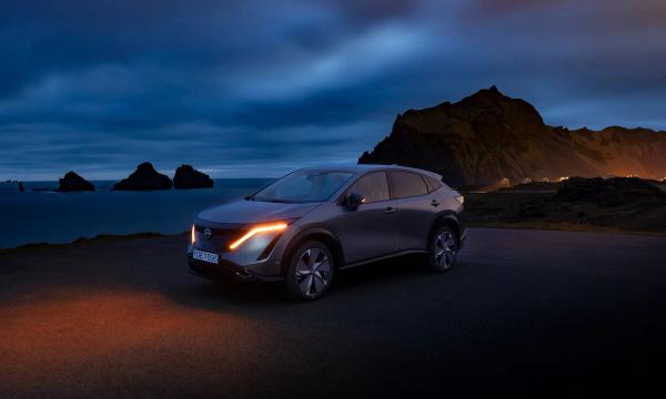 A grey Nissan Ariya with amber lights on, parked on a dark coastal road with mountains in the background.