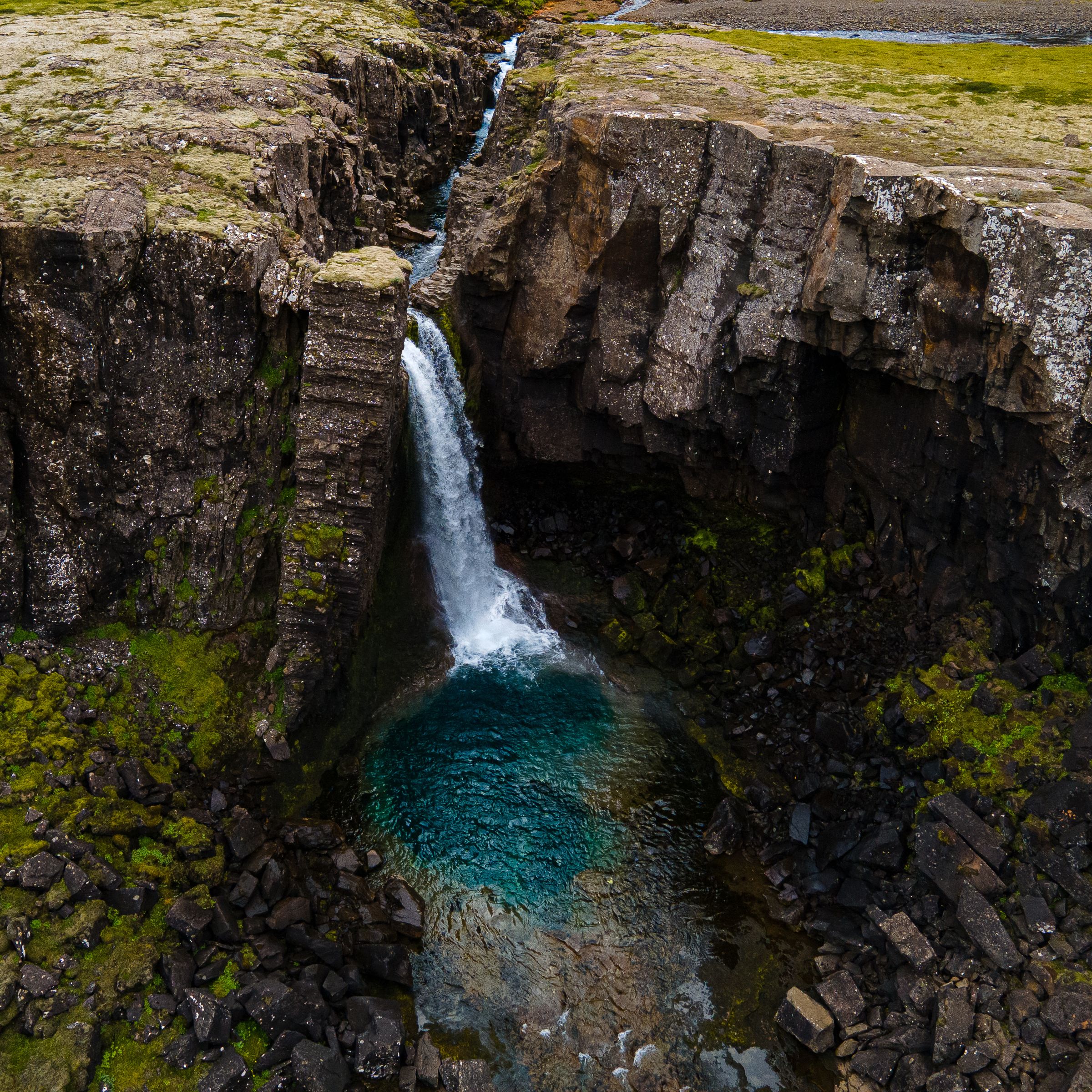 A waterfall plunges into a vibrant blue pool, surrounded by dark, mossy rock cliffs.