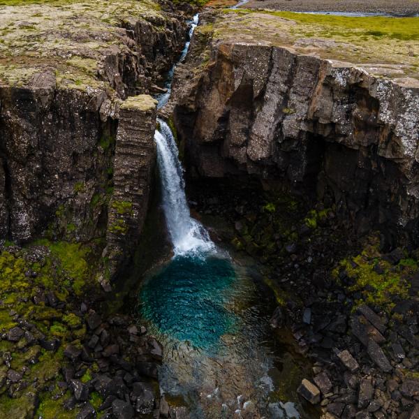 A waterfall plunges into a vibrant blue pool, surrounded by dark, mossy rock cliffs.