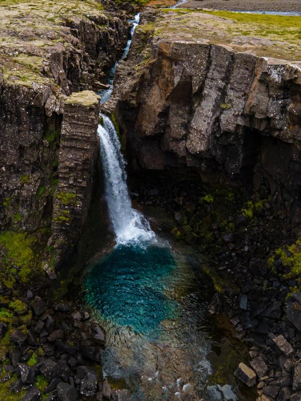 A waterfall cascades into a vibrant blue pool within a rugged, mossy canyon.