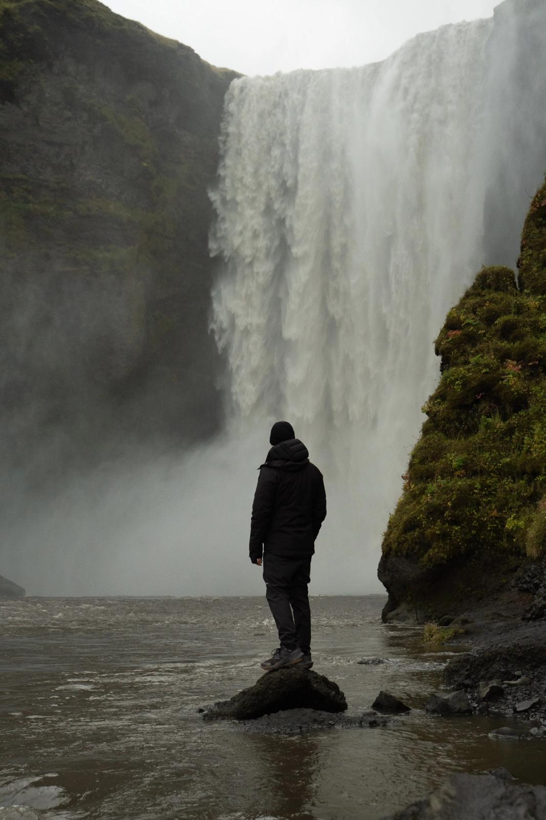 A person in a dark jacket stands on a rock in a river, facing a tall waterfall.