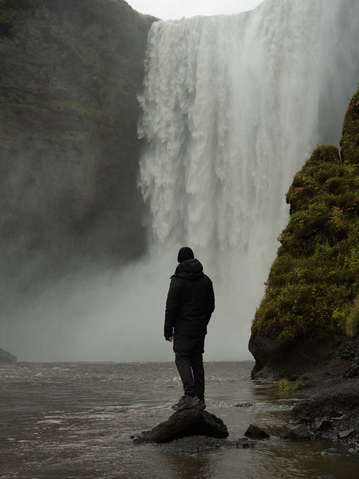 A person in a dark jacket stands on a rock in a river, facing a tall waterfall.