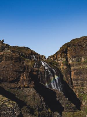 Una cascada en varios niveles desciende por oscuros acantilados rocosos bajo un cielo azul despejado, mientras un arcoíris se forma entre sus aguas.