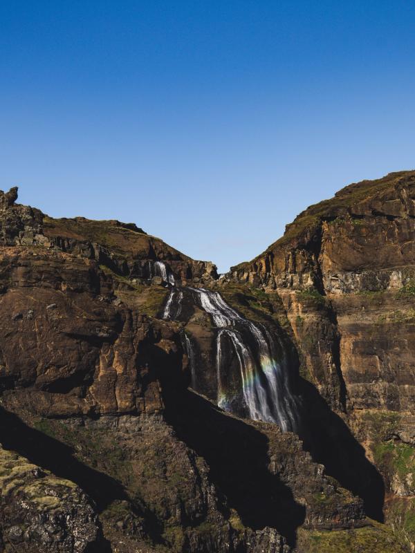 A tiered waterfall with a rainbow cascades down dark, rocky cliffs under a clear blue sky.
