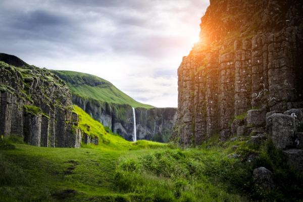 Green valley with basalt columns, a distant waterfall, and bright sun flare on the right.