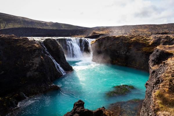 A powerful waterfall cascades into a vibrant turquoise river surrounded by dark, rugged cliffs.