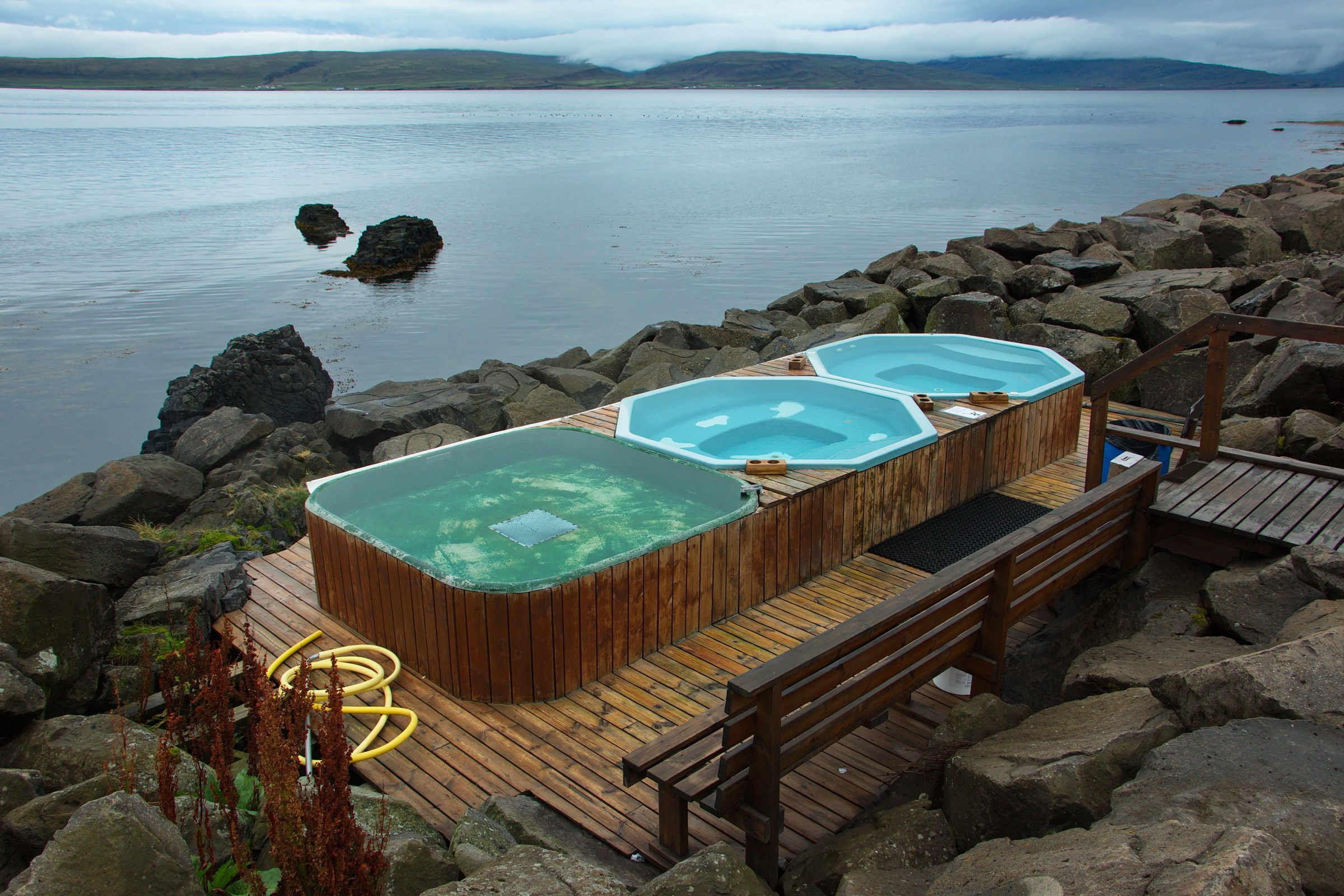 Three outdoor hot tubs on a wooden deck next to a rocky shore, overlooking a calm bay with distant hills.