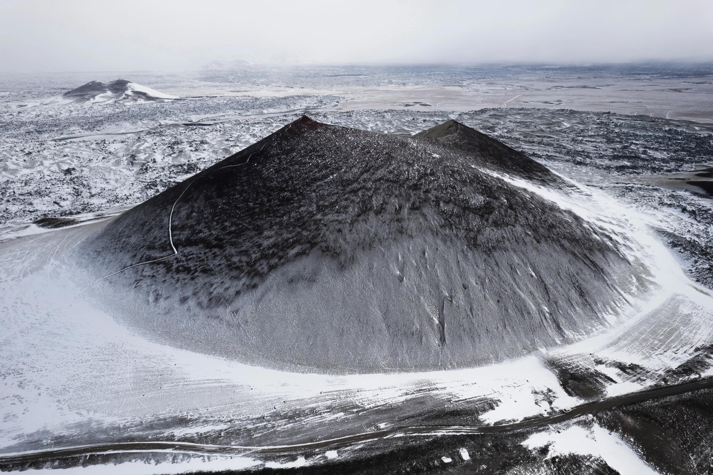 Dark volcanic cone partially covered in snow, surrounded by a snowy, desolate landscape.