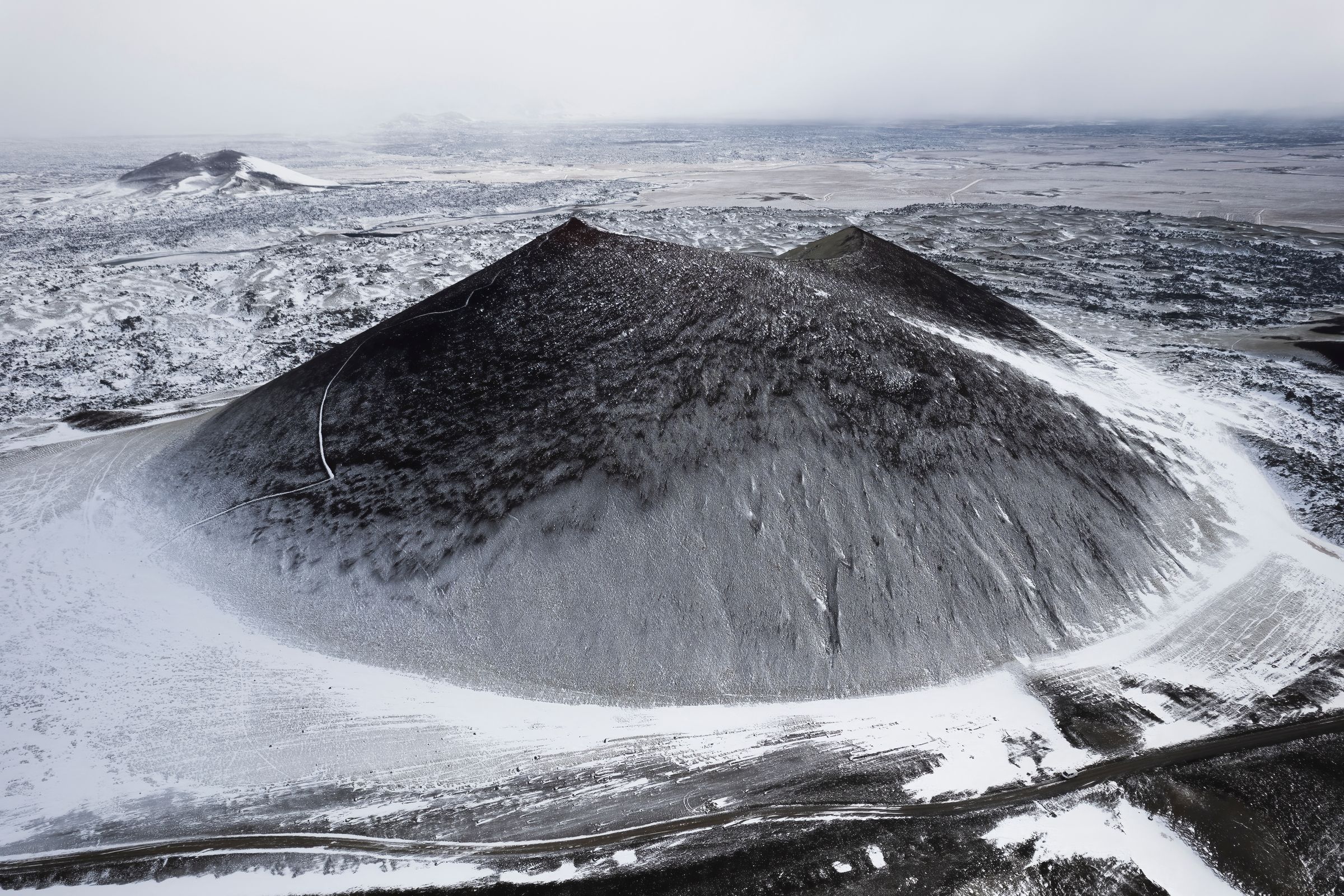 Dark volcanic cone partially covered in snow, surrounded by a snowy, desolate landscape.