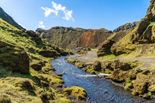 A clear river flows through a green, rocky valley with moss-covered slopes under a blue sky. Distant buildings are visible near a mountain with a reddish-brown section.