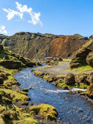 A clear river flows through a green, rocky valley with moss-covered slopes under a blue sky. Distant buildings are visible near a mountain with a reddish-brown section.