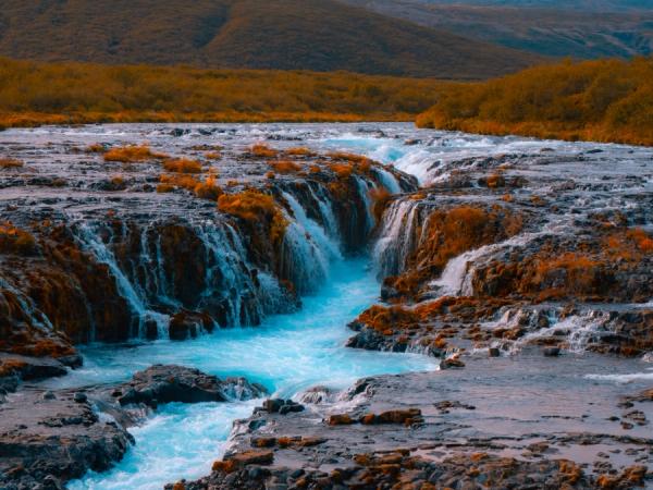 A bright blue river cascading over rocks with reddish-orange foliage on the banks.