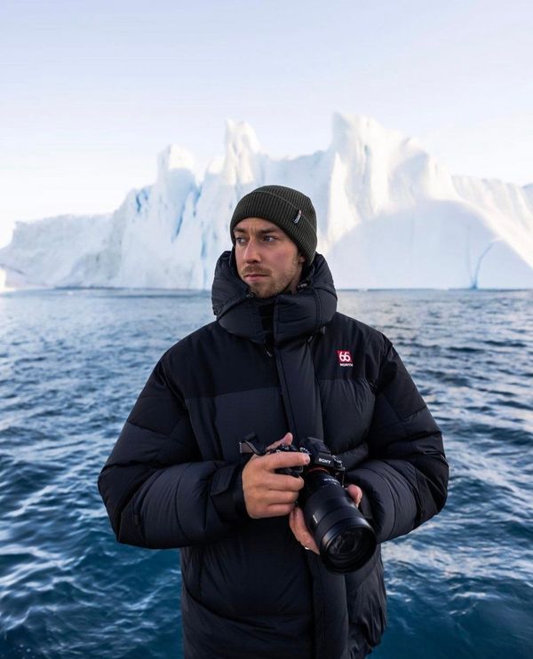a man in a black jacket is holding a camera in front of a large iceberg .