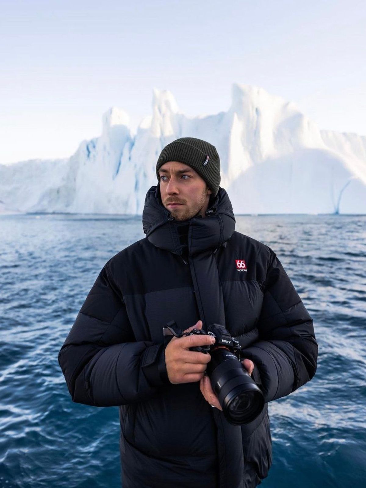 a man in a black jacket is holding a camera in front of a large iceberg .