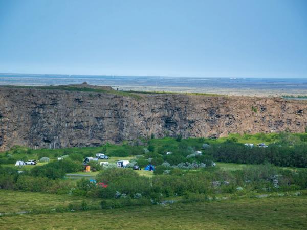 A campsite with tents and RVs sits at the base of a tall cliff, topped by a flat, wide landscape under a blue sky.