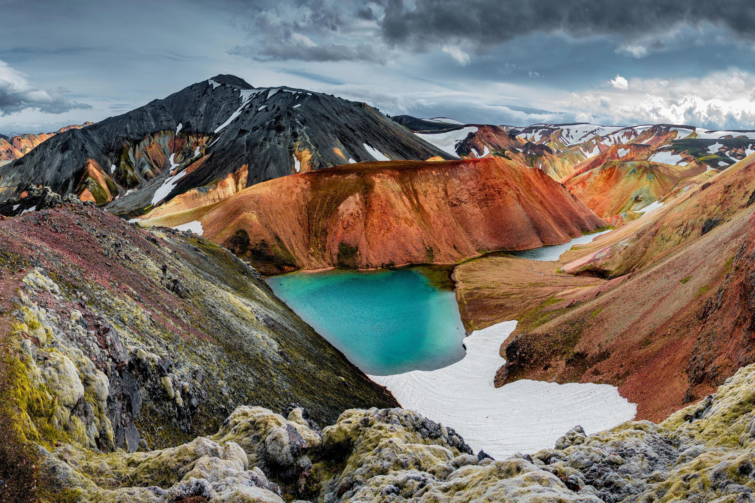 A vibrant turquoise lake surrounded by colorful, reddish mountains with snow and dark peaks under a cloudy sky.