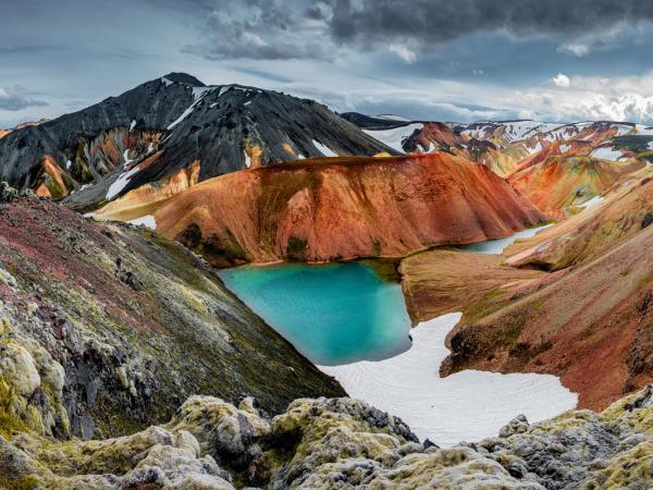 A vibrant turquoise lake surrounded by colorful, reddish mountains with snow and dark peaks under a cloudy sky.