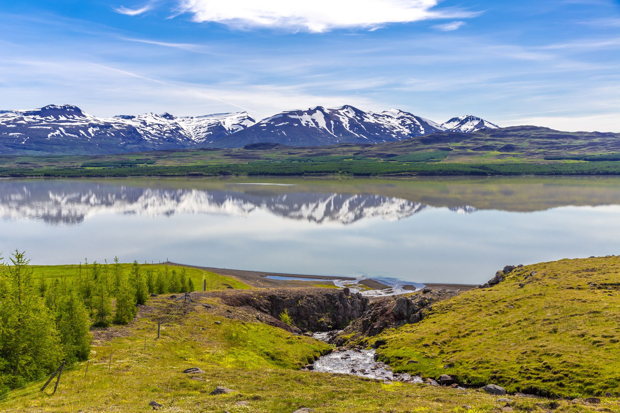 Snow-capped mountains reflect in a calm lake under a blue sky, with green hills and a stream in the foreground.