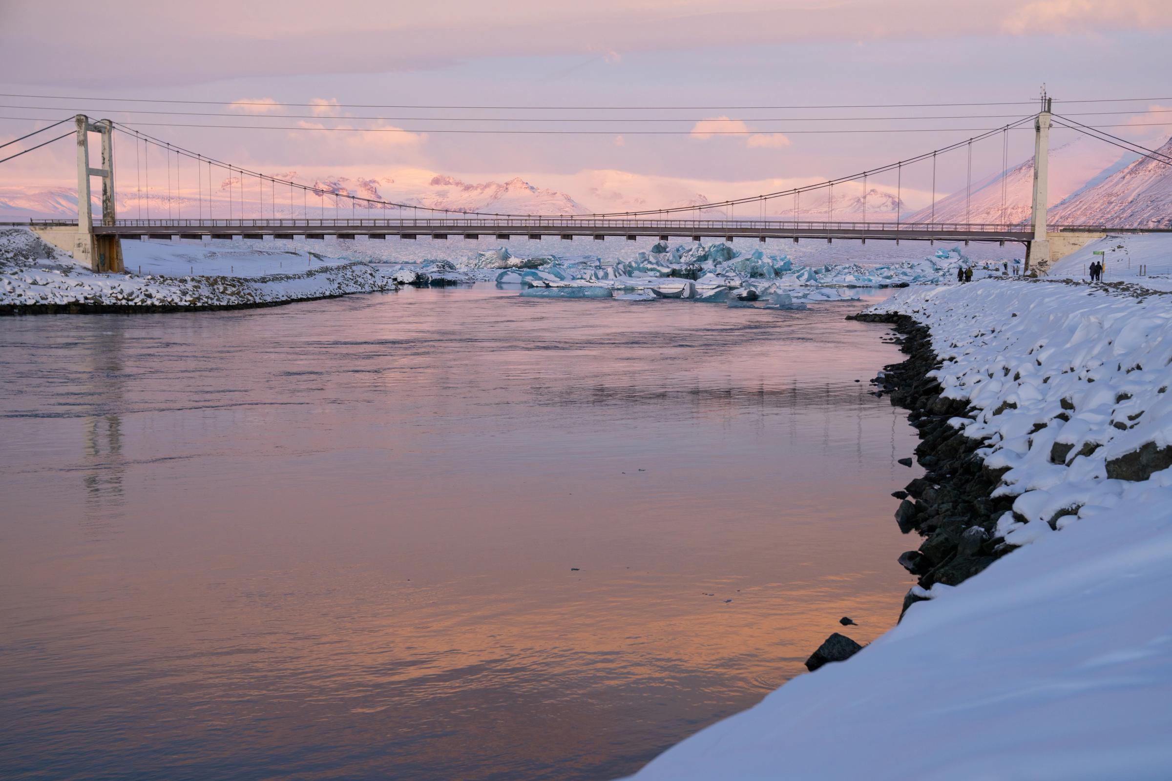 Un puente colgante cruza un río que refleja un cielo rosado, con icebergs en una laguna y montañas nevadas al fondo.
