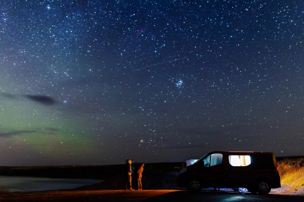 Dos personas con un telescopio y una furgoneta camper bajo un brillante cielo estrellado con una tenue aurora.