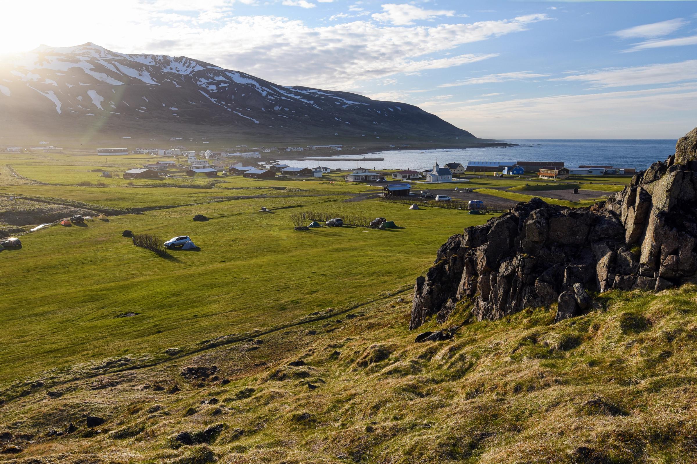A bright, coastal landscape with a green valley, campers, a village by the sea, and snow-capped mountains.