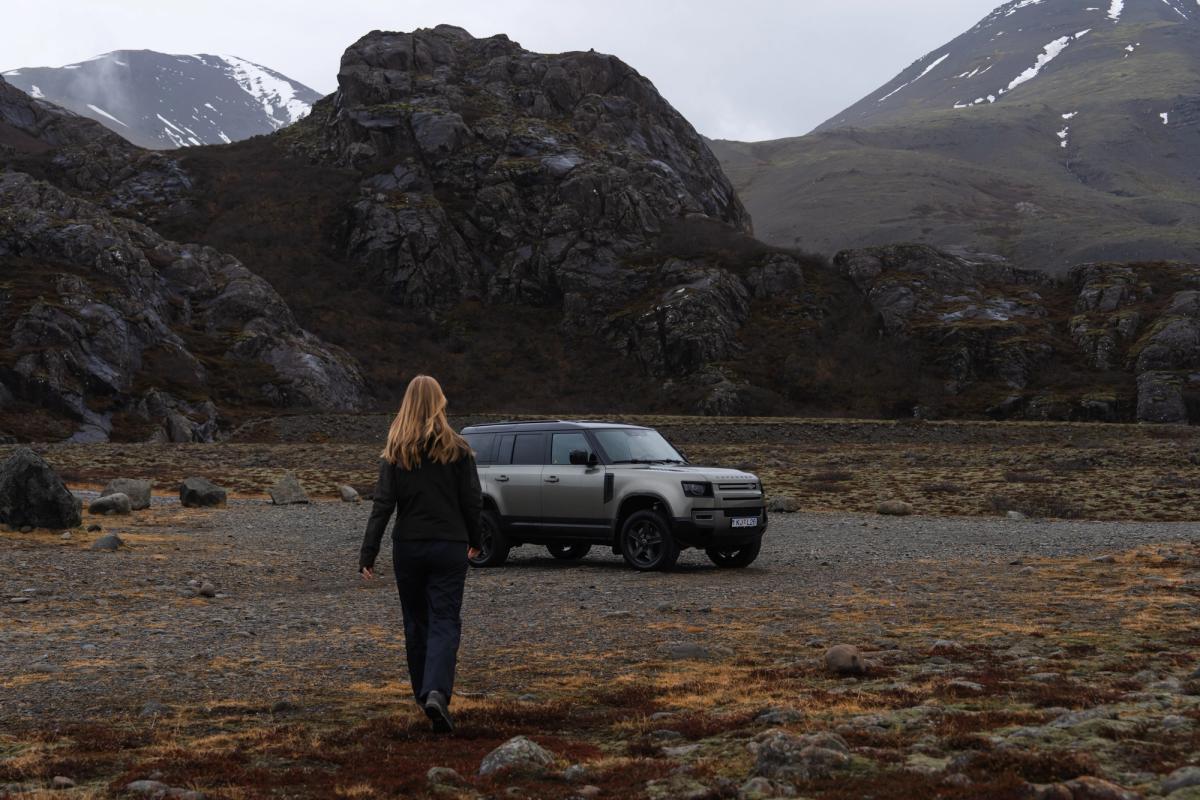 a woman is walking towards a land rover defender parked in a field .