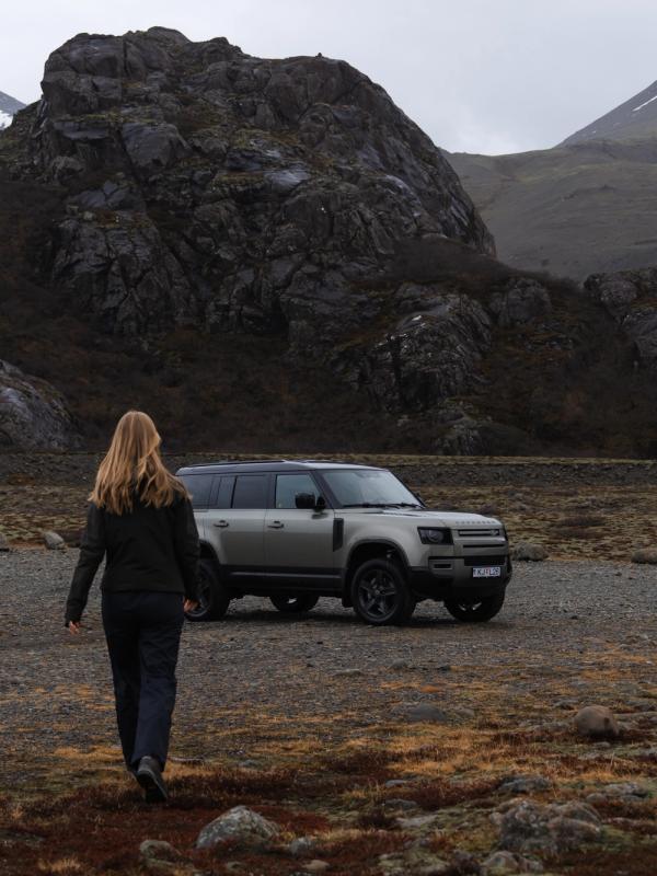 a woman is walking towards a land rover defender parked in a field .