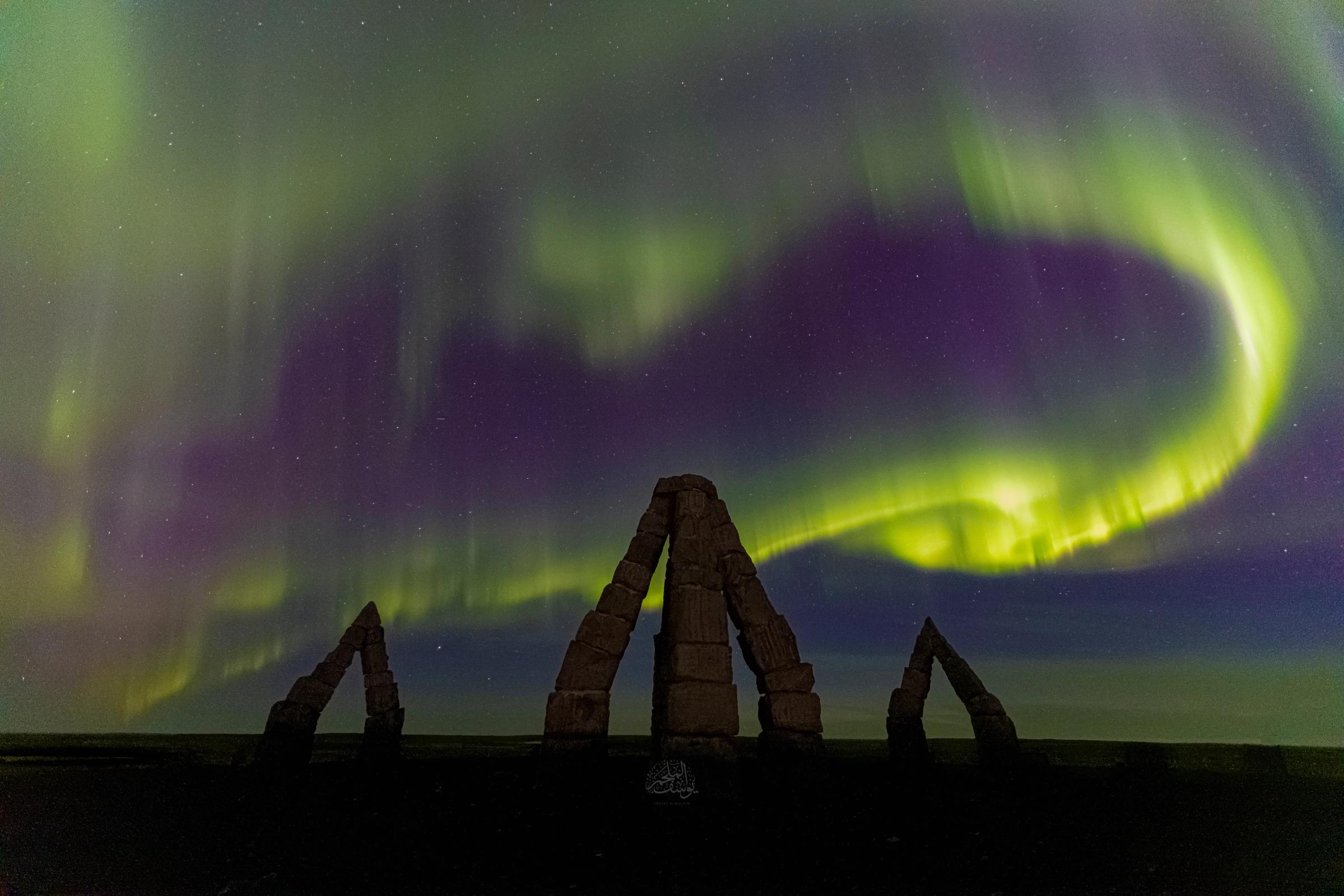 Auroras boreales verdes y moradas se arremolinan sobre tres monumentos de piedra oscuros.