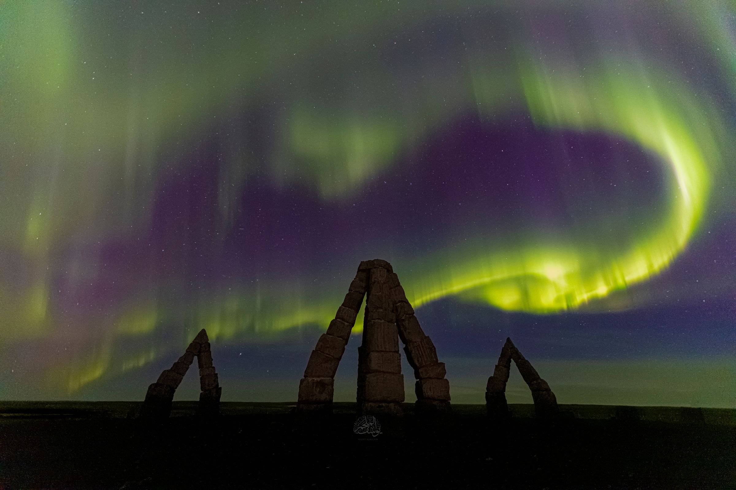 Auroras boreales verdes y moradas se arremolinan sobre tres monumentos de piedra oscuros.